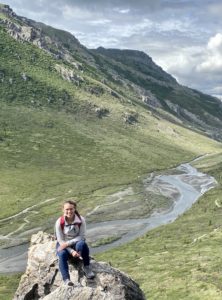 Young woman sits on rock overlooking a gray-blue river and green mountain valley