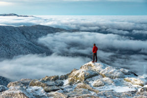 Hiker in red jacket stands on a rock outcropping above an undercast cloud layer
