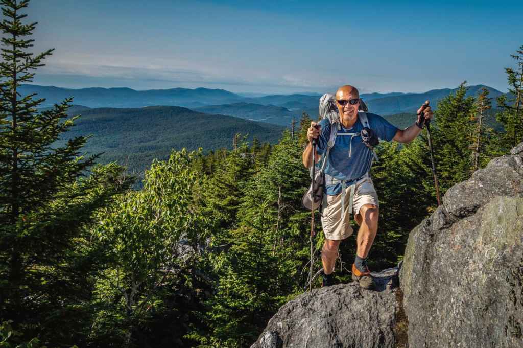 Man climbs rocky Mt Mansfield with landscape behind him.