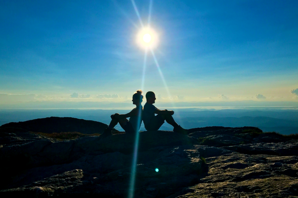 Two hikers sit back-to-back with the sun behind them atop Mt. Mansfield. 