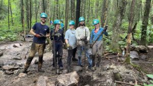 5 young adults wearing hard hats and muddly clothes pose for camera on muddy trail