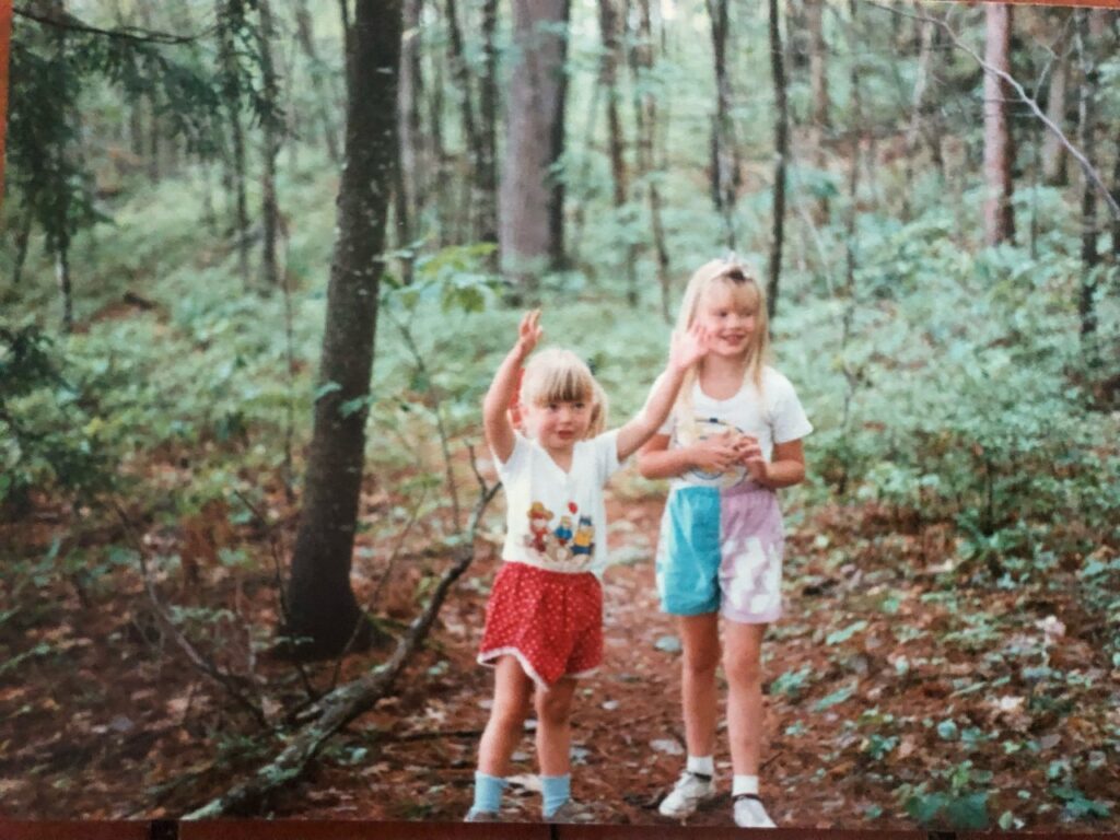 the author, right, and her sister as young kids, hiking. The left sister has red shorts and has her hands up in the air. Both girls have white blond hair and white t shirts.