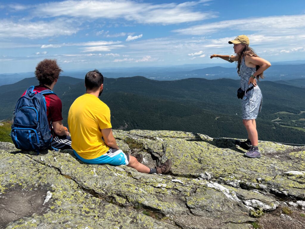 caretaker Kate songer, at right, stands on the summit of a mountain pointing off into the distance. Two hikers, one with a blue backpack and one in a yellow shirt, sit on the rocks on the left side of the image. All persons are facing away from the camera. 