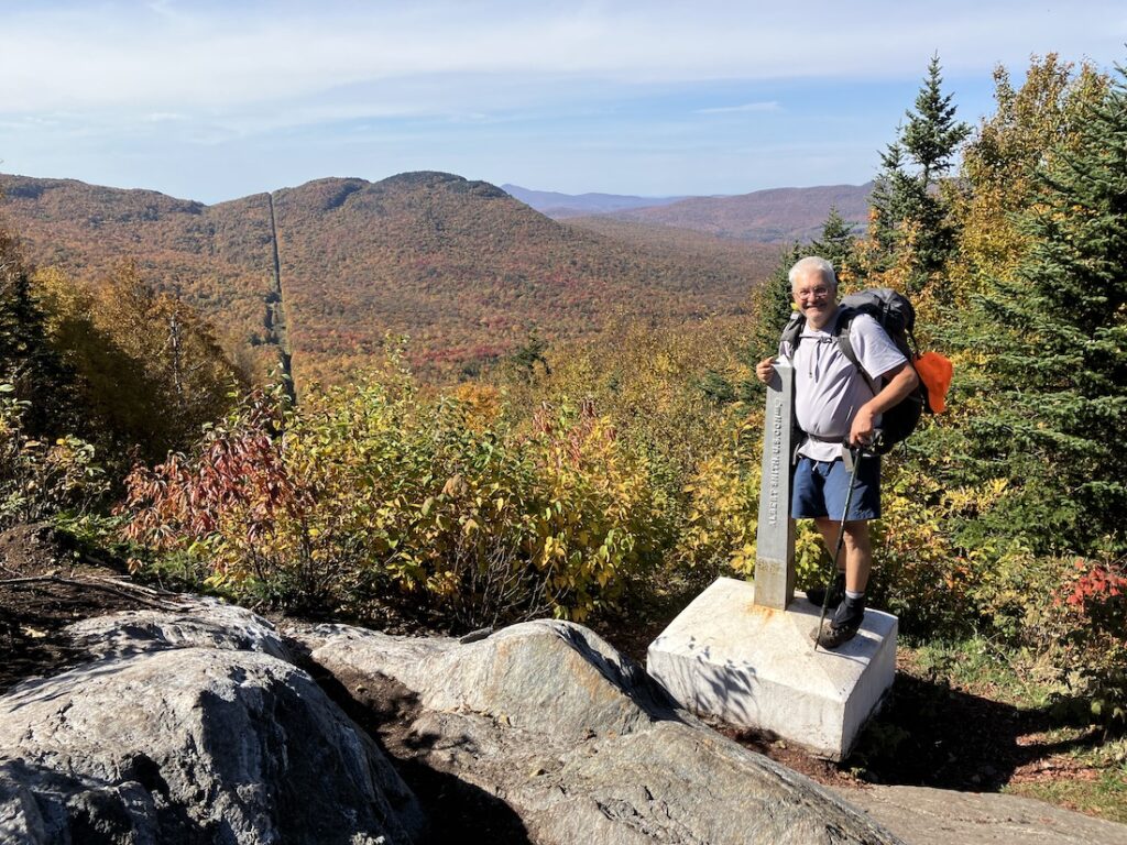 an older man with a red pack poses on the right side of the journey's end monument