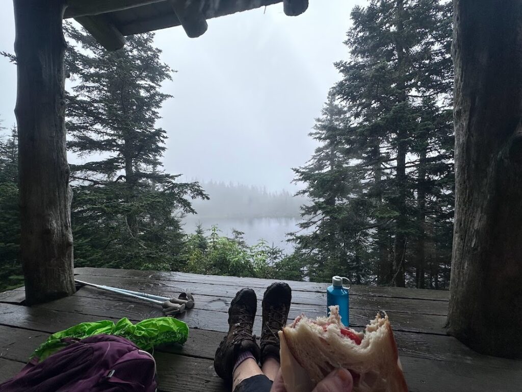 hikers feet, trekking poles, water bottle, and sandwich in the flood of a shelter, looking out over a rainy cloudy pond