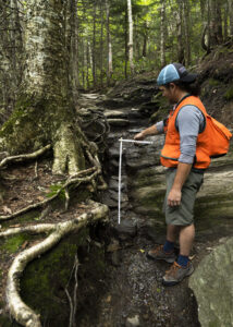 Keegan, in orange survey vest, uses a tape measure to measure erosion on the sterling pond trail