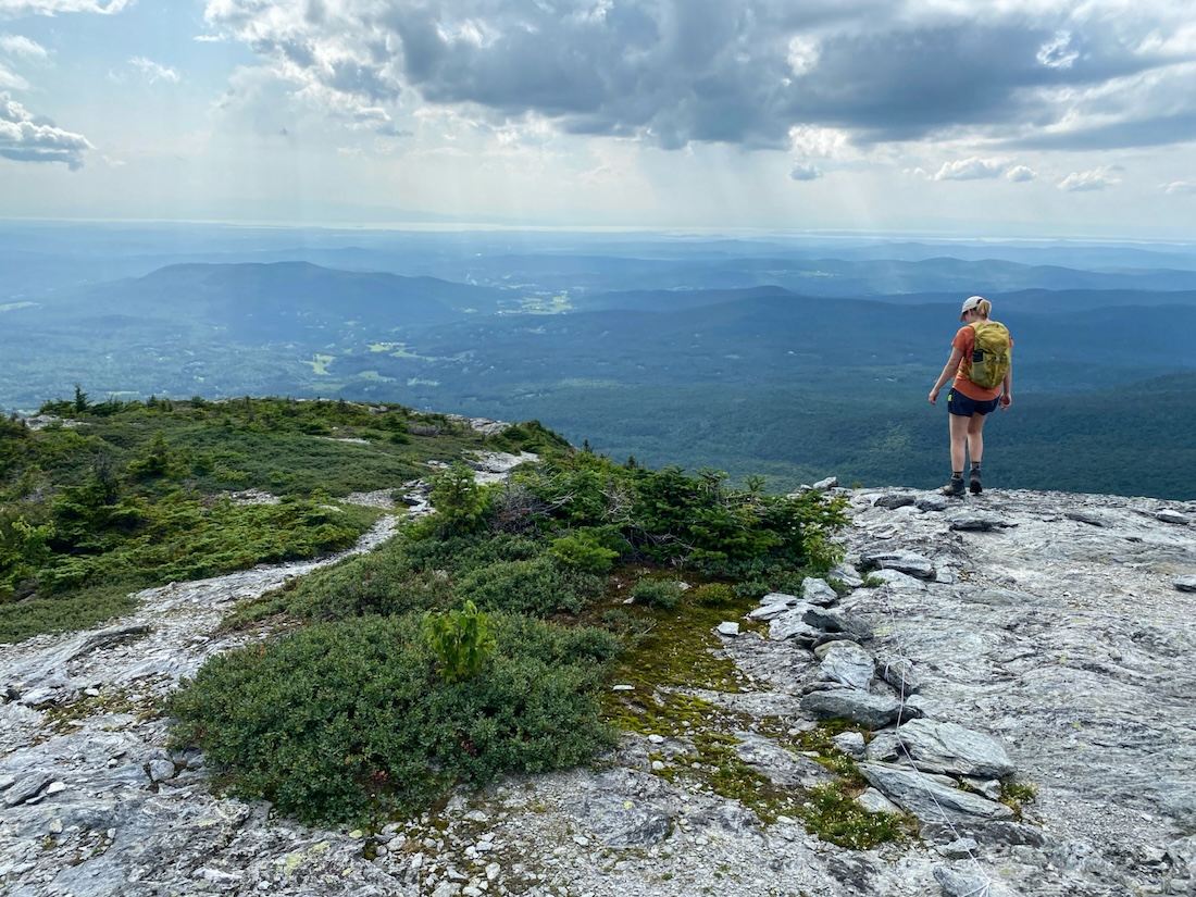 a hiker stands on a rocky summit overlooking the view below. Rocks and green vegetation are in the foreground