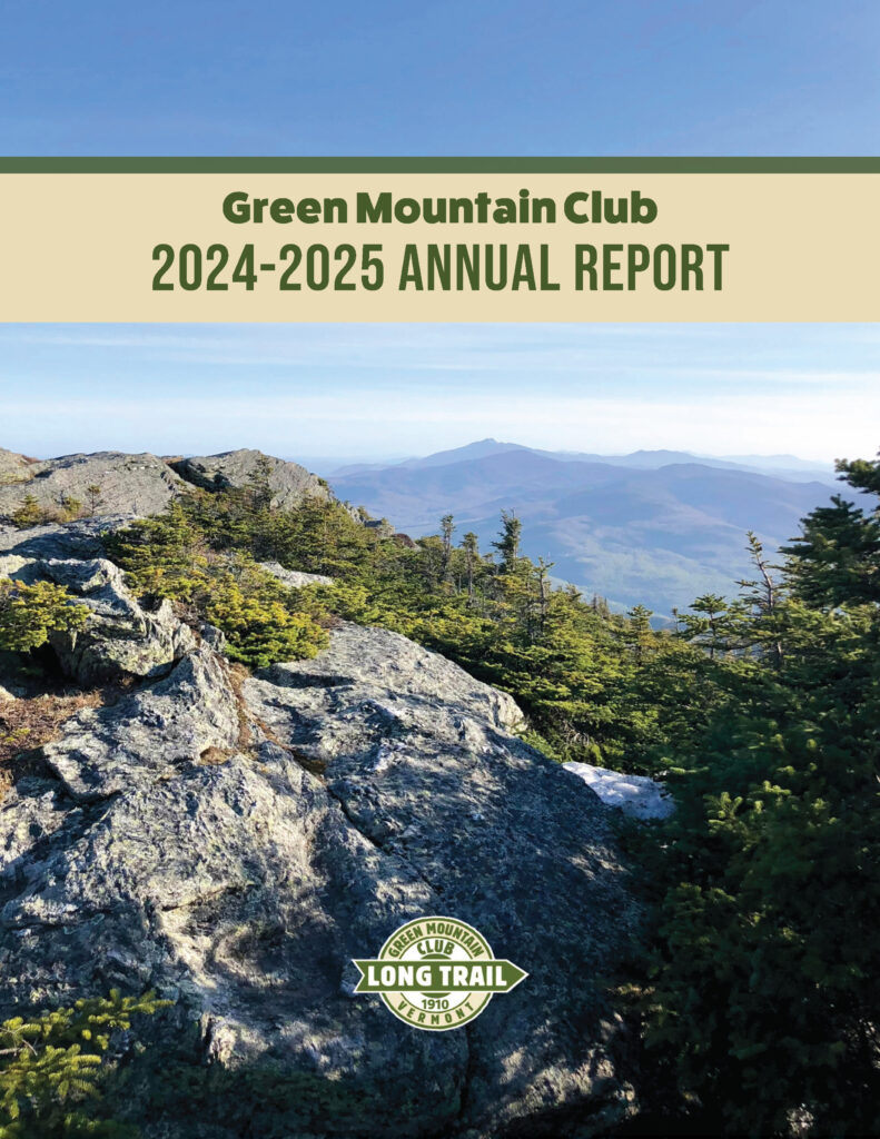 a scenic image of a rocky summit in the foreground, some shrub greenery in the middle gorund, and blue sky and distant mountains in the background