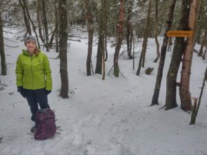 a woman in a green jacket stands in front of a snowy trail flanked by trees